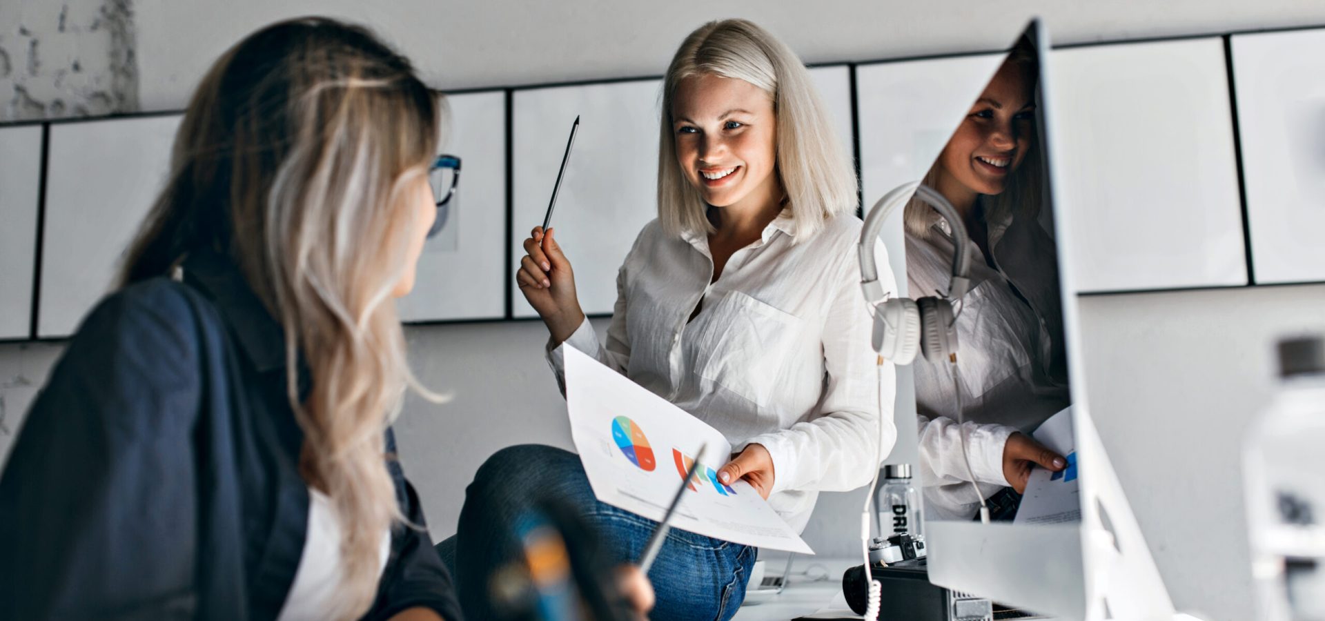 Smiling blonde female manager holding infographic and pencil, while sitting on table. Indoor portrait of two girls working with computer in office.
